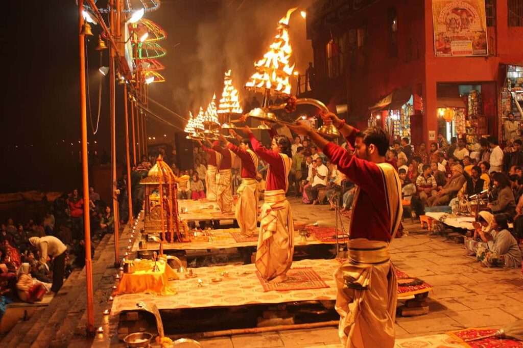 Hindu priests performing Ganga Aarti ceremony at the river Ganges in Varanasi, India, while people gather to pray and offer flowers and candles."