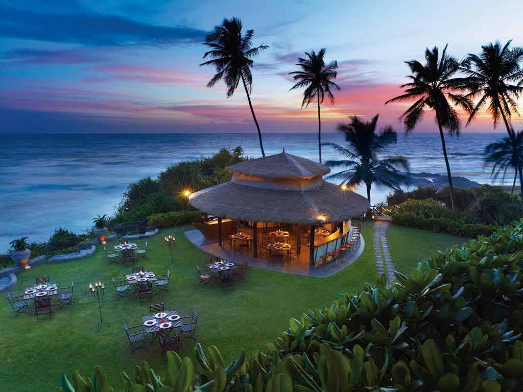 A view of the Shangri-La Hambantota Golf Resort & Spa located on a pristine beach in Sri Lanka, with palm trees, blue sky, and turquoise water in the background. The image shows the resort's luxurious outdoor pool and sun loungers.