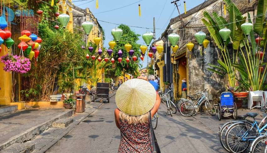 "A woman wearing a Vietnam cap stands on a street decorated with hanging lanterns."