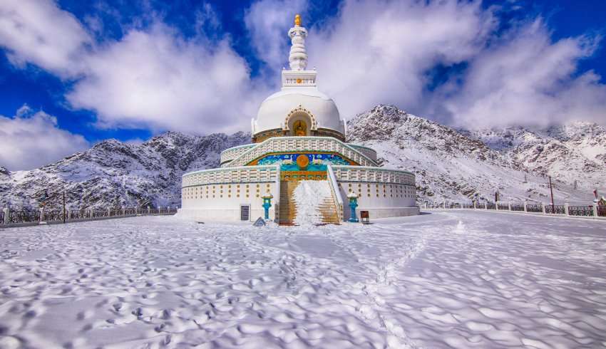 "Shanti Stupa, Ladakh surrounded by snow-capped mountains and covered in snow."